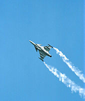 Jet aircraft performing rolls while climbing, revealing its underside, where missiles are attached. White smoke trail each wing tips.