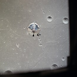 The top of the silvery command module is seen over a grey, cratered lunar surface