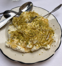 A plate of ashta, a dairy dessert topped with crushed pistachios and honey, with three spoons.