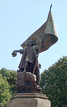 公園に設置されている大きな像の写真。髭をたくわえたロングコート姿の男性が岩の上に立っており、右手に帽子、左手に大きな旗を持っているデザイン。