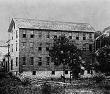 Grainy photo that was published in an old newspaper and then scanned in modern times. It shows a large rectangular building with a stone ground level, then three floors above that, and a pitched roof. Each floor has 10 windows along the longest side of the building, and 2 windows on the shorter sides, and some of the windows are open. Several trees are near the building, and a split fence encircles the area.