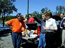Sekiz orta yaşlı insandan oluşan bir grup, turuncu trafik konileri ve beyaz yiyecek kapları taşıyan bir kamyonetin açık arkasını çevreliyor.  Sahne, mavi gökyüzü güneşli bir günde, bir su kütlesinin, bazı ağaçların ve bir telefon direğinin yanındadır.  Üç kişi en belirgindir.  Bir adam mavi pantolon ve tehlikeli maddeler ekibinden turuncu tişört ve mavi ve turuncu beyzbol şapkası giymiş.  Başka bir adam görünmeyen bir nesneyi işaret ediyor ve kot pantolon ve lacivert bir gömlek giyiyor ve onun hakkında bir otorite havası var.  Bir kadın o adamın yanında duruyor, dinliyor.  Siyah pantolon, beyaz bir gömlek giyiyor ve kulağının arkasından yukarısı ve aşağısı kesilen kahverengi saçları var ve ince siyah bir kol saati takıyor.