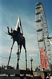 Exposition de Dalí à Londres. Au premier plan, une sculpture d'éléphant aux pattes arachnéennes, avec une pyramide sur le dos semblable à celles qui apparaissent sur la toile. Au fond se trouve London Eye.