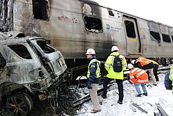Several people in neon colored safety clothing and white helmets examine the fire-damaged wreckage of a silvery passenger rail car along the top with "Metro-North Railroad" written on it. One person has the letters "NTSB" on their jacket sleeve. At left is the rear of a similarly fire damaged automobile.