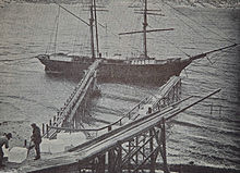 Photographie de deux hommes devant plusieurs blocs de glace sur un ponton en bois au bord de la mer. Plusieurs rampes en bois sur pilotis s'avancent au-dessus de la mer et un navire à deux mats est ancré à l'extrémité de l'un d'eux.