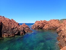 Cliché numérique couleur. Sur un ciel bleu se détachent des roches rouges plongeant dans la mer.