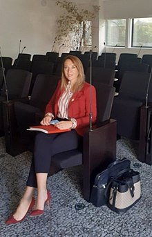 Stéphanie Galzy, age 41. She is sat on a row of seats with a writing book and she is wearing a red jacket and a white shirt with red pinstripes, she is also wearing red heals