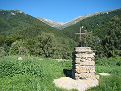 Photographie d'une vallée pyrénéenne, arborée au second plan et dénudée en arrière-plan plus en altitude. Au premier plan, une croix de fer.