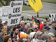 Een groep mensen bij een straathoek op de achtergrond, van bovenaf gezien.  Links houdt men een zwart-op-wit bord omhoog met de tekst "Obama Lies, Grandma Dies".  Delen van andere tekens zijn zichtbaar en een deel van een Gadsden-vlag hangt van boven de bovenkant van de afbeelding