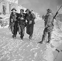 two men each side of a wounded man walking along a snow covered road being watched by rifle armed sentries