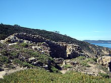 Farbfoto von Ruinen in der Garigue.  Das Meer scheint überwältigt.