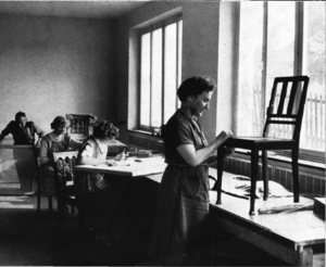 Werkstatt mit Flechtarbeiten, eine Frau stehend an einem Stuhl arbeitend, im Hintergrund weitere Personen sitzend und stehend. Schwarzweißfoto aus dem Jahr 1956