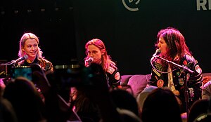 Phoebe Bridgers, Julien Baker, and Lucy Dacus sit at microphones in front of an audience. Bridgers is smiling slightly while she and Dacus are looking at each other past Baker, who is blankly looking straight ahead.