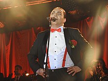 A Man with black-dyed hair is wearing a tuxedo with a red bowtie with his hands on his hips