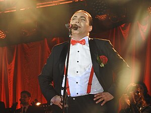 A Man with black-dyed hair is wearing a tuxedo with a red bowtie with his hands on his hips