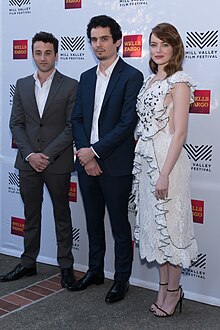 Justin Hurwitz og Damien Chazelle i dress og Emma Stone i en hvit kjole med sorte prikker.  De to mennene krysser hendene mens Emma Stone er i profil med hodet vendt mot oss.  Hurwitz stirrer på bakken mens Chazelle og Stone ser på linsen.