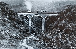 En stein viadukten med tre buer som spenner over en bratt ensidig kløft. Et damptog står på viadukten, med et lokomotiv, en godsvogn og tre passasjervogner.