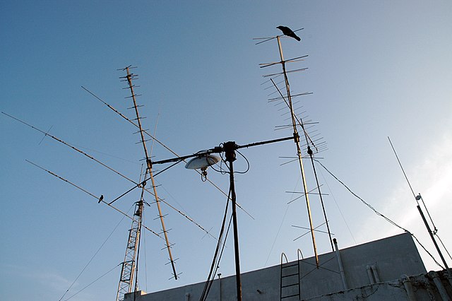 Thin, black, straight wires intersecting at various points protruding from a grey building or else from each other with a blue sky in the background