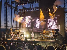 A brightly lit concert stage at sunset with puffs of smoke, television screens on the backdrop, and an audience watching