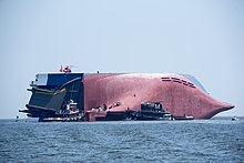 Remote view of a large ship listing 90°, its red hull largely out of the water, with two large tugboats and several smaller boats attending to it