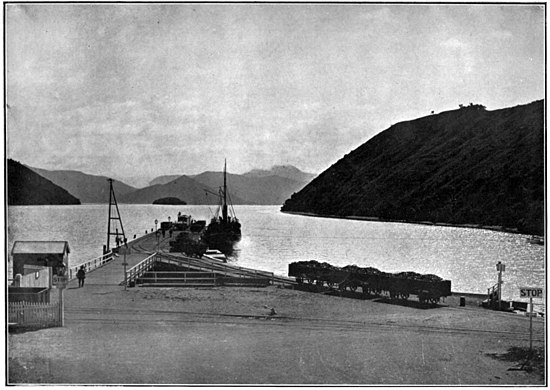 Wharf with a sailing ship alongside, with train tracks and wagons, and a calm sea surrounded by hills either side and in the distance