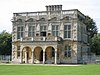 A five-bay, flat-roofed building with two storeys and an arched single-storey portico. Surrounding the top of the building is a balustrade and chimneys rise from the rear