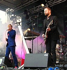 Двое мужчин стоят на сцене с микрофонами, пока один играет на гитаре