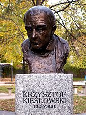Buste de Kieślowski de couleur bronze. Il est posé sur une colonne de marbre qui indique le nom du réalisateur et le titre Rezyser.