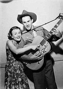 Une image trois quarts en noir et blanc d'un couple.  La femme à gauche porte une robe à fleurs et a sa main droite posée sur la corde de la guitare.  Elle est tenue par l'homme de vingt-cinq ans qui porte une chemise de cow-boy et un chapeau.  Sa main gauche est sur le manche et sa droite est en dessous de la main de la femme.