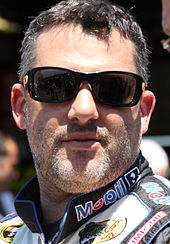 A man in his early forties sporting a stubble and wearing black sunglasses