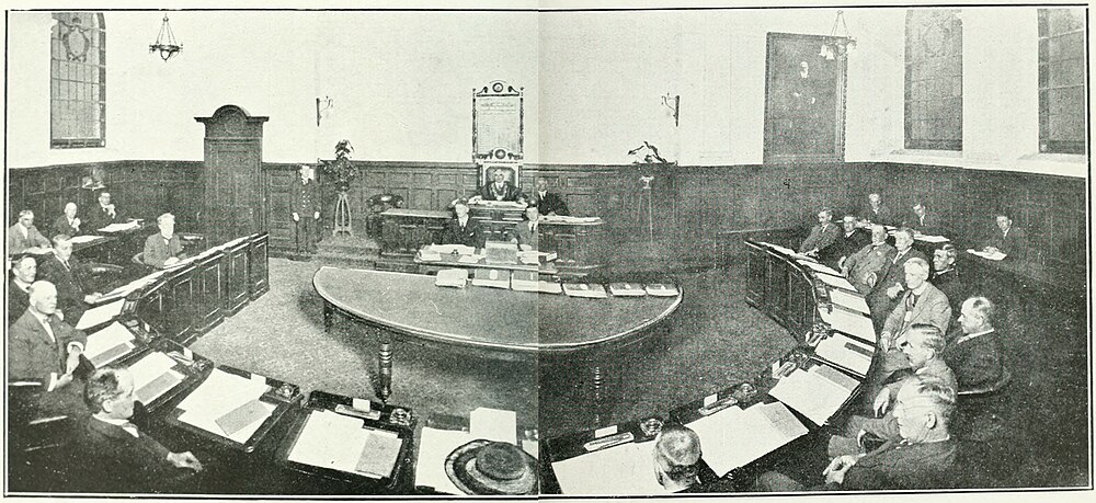 City Councillors sitting behind tables in a semi-circle, with the Mayor on a raised desk in the center, and other council staff city seated before the Mayor and a half round table with papers on it.