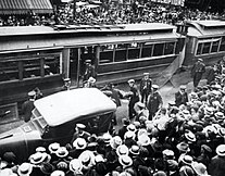 Two people carry an injured man on a stretcher, nearby a trolley, to an ambulance while a crowd onlooks. Denver Streetcar Strike, Man carried on stretcher.jpg