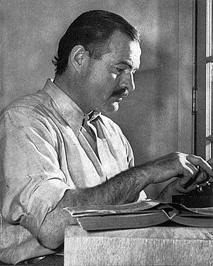 Dark-haired man in light colored short-sleeved shirt working on a typewriter at a table on which sits an open book