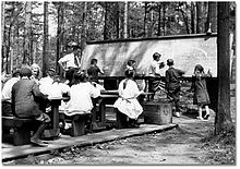 group of children in an outdoor classroom seated at desks and standing at blackboard