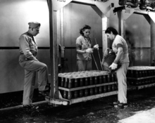 Photograph of a man in a military uniform observing two young women working at a rack of mortar shells