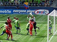 Vários jogadores com uniformes vermelhos e verdes na frente do gol com um goleiro de preto pulando com a mão batendo em uma bola de futebol