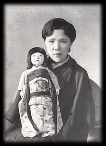 <Black and white portrait photograph of a woman with short, dark hair, wearing a dark dress, holding a doll>