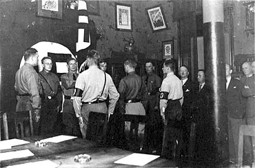 Black-and-white photograph of Nazis standing in a decorated office. An empty table is in the foreground. Three men look towards the camera, including one in the middle with a toothbrush moustache.