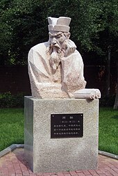 Photographie montrant la sculpture d'un buste de Shang Yang en marbre dans un jardin. L'homme porte un bouc et un chapeau. Il tourne la tête vers la droite, a sa main gauche sur sa joue gauche et tient un manuscrit dans sa main droite.