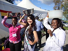 A photograph of three smiling women standing in a row while balancing bowls on their heads, all in front of a white tent under a blue sky