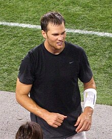 A man standing on the sidelines of an American football field. He is wearing a black shirt.