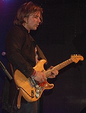 Lukas Nelson hraje na kytaru v černém tričku a šátku.
