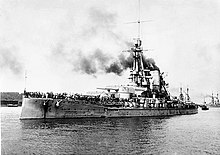 A large warship steams at low speed; gray smoke drifts from the smoke stack