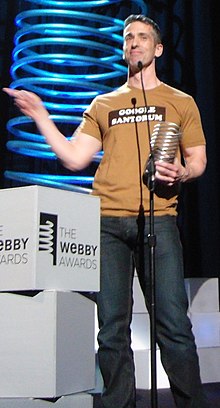 Dan Savage at the Webby Awards, holding his award and wearing a t-shirt that reads “GOOGLE SANTORUM”.