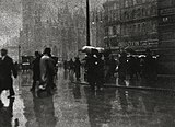 Black and white photo of a street corner in Vienna. Street wet with rain and people walking in various directions some people carrying umbrellas.