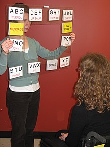 Un utilisateur en chaise roulante fait face à un interlocuteur qui tient un tableau en matière plastique transparente entre eux, les lettres de l'alphabet et des messages sont imprimés sur des cartons disposés sur le pourtour du tableau