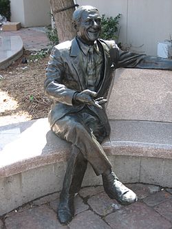 Une statue assise sur un banc incurvé, derrière lequel se trouve le tronc d'un arbre adjacent au mur d'un bâtiment.