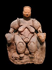 Statuette of a nude, corpulent, seated woman flanked by two felines from Catalhoyuk, dating to c. 6000 BCE, thought by most archaeologists to represent a goddess of some kind Seated Woman of Catalhoyuk on black background.jpg