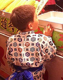 Мальчик стоит у прилавка с кукурузой в початках спиной к зрителю;  две вертикальные складки, направленные наружу, спускаются с его плеч.