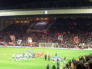 A stand which is full of people set behind a field of grass. There are a number of flags and banners in the crowd. On the field of grass are a number of people.